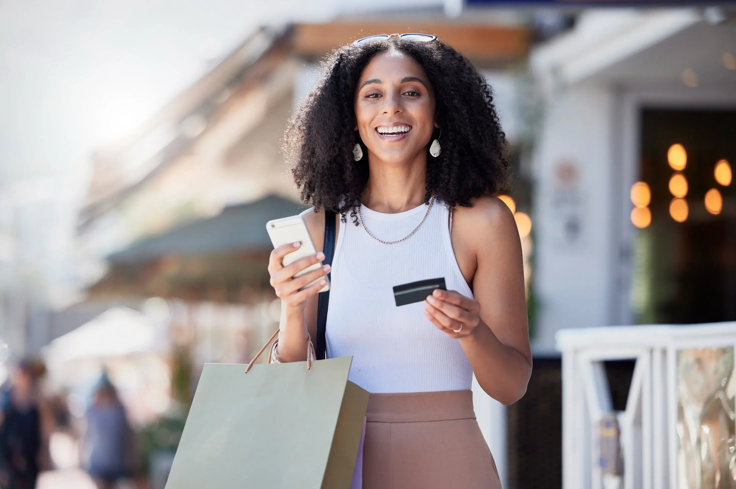 Happy young woman with curly hair holding a credit card and smartphone while standing outdoors with a shopping bag.